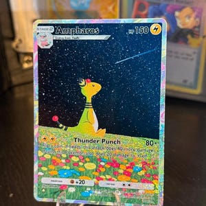 May include: Ampharos Pokémon trading card with a starry night background. The card features a yellow and green Ampharos character with the text "Thunder Punch" and "HP 150". The card is displayed in a clear stand.