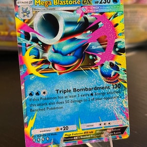 Puede incluir: Una carta coleccionable Pokémon Mega Blastoise EX. La carta presenta una ilustración de Blastoise azul y blanco con detalles en rosa y amarillo. La carta tiene el texto "Triple Bombardment 130" y "HP 230".