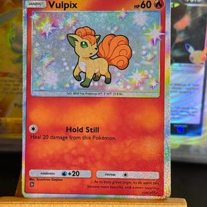 May include: A holographic Vulpix trading card featuring a tan fox-like creature with a curly orange tail. The card displays 60 HP, the move Hold Still, and text detailing its height of 0.6 metres and weight of 9.9 kilograms, set against a shimmering rainbow background.