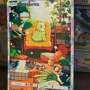 Op de afbeelding: Een holografische Pokémon-verzamelkaart met het personage Chickorita in een gezellige kamer. De kaart heeft een groen en geel kleurenschema, met de tekst "Razor Leaf" en "+20" zichtbaar. De kaart wordt op een transparante standaard gepresenteerd.