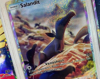 May include: A holographic Pokemon trading card featuring Salandit, a small, gray, lizard-like Pokemon with red eyes. The card has a background of a rocky, desert-like landscape. The card text reads "Venoshock 10+ If your opponent's Active Pokemon is Poisoned, this attack does 40 more damage."
