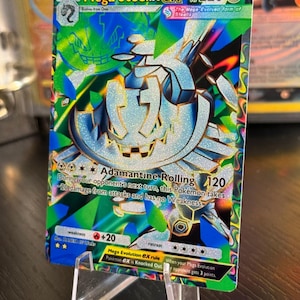 May include: A holographic Mega Steelix EX Pokémon card. The card features a metallic silver and blue Steelix design with a green and blue background. The card is displayed on a clear plastic stand.