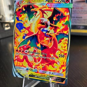 May include: A holographic Mega Charizard Y EX Pokémon trading card. The card features a fiery orange and red Charizard with blue accents, set against a background of flames. The card has a HP of 220 and a Crimson Dive attack.
