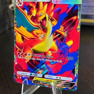 Könnte beinhalten: Eine Mega Charizard Y EX Pokémon-Karte mit einer lebendigen Illustration von Charizard in Rot-, Orange- und Blautönen. Die Karte zeigt den Text "Crimson Dive" und "HP 220". Sie wird auf einem transparenten Acrylständer präsentiert.