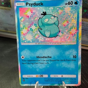 Puede incluir: Una carta coleccionable Pokémon holográfica de Psyduck. La carta presenta un personaje Psyduck azul con un mechón de pelo negro. La carta tiene un borde azul y el texto "Psyduck" y "HP 60".