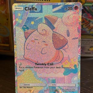 May include: A holographic Cleffa Pokémon card with a pink and blue design. The card features the text "Cleffa" and "Twinkly Call". The card is displayed in a clear plastic holder.