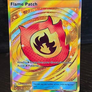 Può includere: Una carta collezionabile dorata con un design a fiamma rossa e arancione. La carta recita "Flame Patch" e include testo sull'attacco di energia ai Pokémon. La carta è tenuta da un supporto di plastica trasparente.