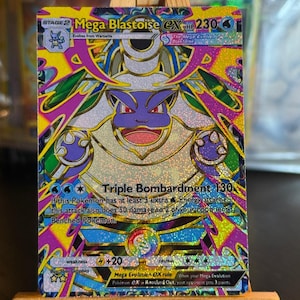 Puede incluir: Una carta coleccionable holográfica Mega Blastoise EX que muestra un personaje morado y azul con cañones en la espalda. La carta presenta 230 HP, el ataque Triple Bombardment y complejos patrones geométricos amarillos, rosas y azules.