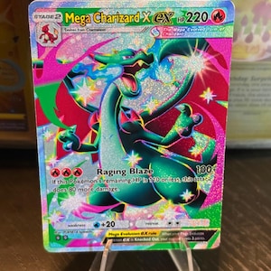 Könnte beinhalten: Eine holografische Mega Charizard X EX Pokémon-Sammelkarte. Die Karte zeigt einen grünen und roten Drachen mit dem Text "Mega Charizard X EX" und "Raging Blaze". Die Karte wird auf einem transparenten Ständer präsentiert.