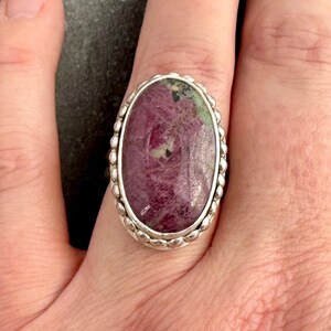 May include: Oval ring with a large, polished ruby gemstone set in silver. The ruby is a deep red colour with green and white inclusions. The silver band of the ring has a beaded design around the gemstone.