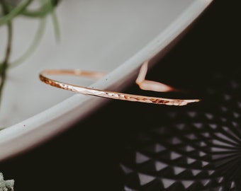 May include: A thin, hammered rose gold bangle bracelet. The bracelet is simple and delicate, with a smooth, polished finish.