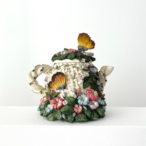 May include: A decorative teapot featuring a white woven basket texture, adorned with sculpted green leaves, pink and blue flowers, and two yellow butterflies. The handle is shaped like a twisted vine, and a butterfly sits atop the lid.