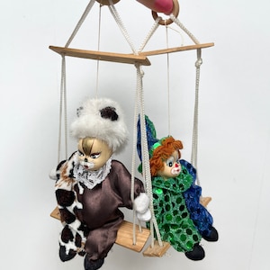 May include: A wooden swing set with two dolls hanging from it. The doll on the left is a brown and white cat with a black nose and whiskers. The doll on the right is a clown with red hair, a green and blue costume, and a red nose.