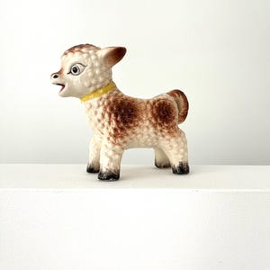 May include: A ceramic lamb figurine with a textured, curly white and brown coat. The lamb has a yellow collar around its neck, large dark eyes, and an open mouth. Its hooves are painted black, and it stands in a profile position facing left.