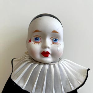 May include: A vintage porcelain doll head with a white face, red lips, and blue eyes. The doll has a black cap, a white ruffled collar, and a red star on its cheek. The doll's face has a tear drop painted on it.