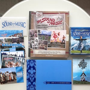 Op de afbeelding: Een verzameling "The Sound of Music"-memorabilia, waaronder een familie-scrapbook, Blu-ray disc, ansichtkaarten en een blauwe doos met een bloemmotief. De items zijn gerangschikt op een wit oppervlak, wat het thema van de film benadrukt.