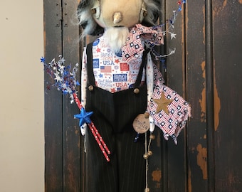 May include: A fabric doll wearing a black hat, black pinstriped overalls, and a white shirt with red, white, and blue patriotic text and flag patterns. The doll holds a star-tipped wand and a small flag-patterned bag with a gold star.
