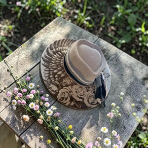 May include: A beige wide-brimmed hat featuring a dark brown printed design of large wings and blooming roses on the brim. The crown is encircled by a black ribbon and a light grey decorative trim, with a dark fabric tie hanging from the side.