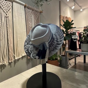 May include: A light gray fedora hat featuring a wide brim decorated with a black and white rose pattern and detailed wing illustrations. The hat is accented with a blue ribbon band, a dark blue fabric tassel, and a delicate white lace trim.