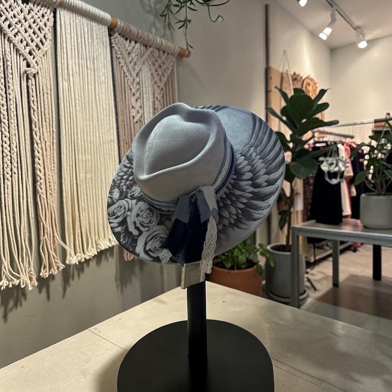 May include: A light gray fedora hat featuring a wide brim decorated with a black and white rose pattern and detailed wing illustrations. The hat is accented with a blue ribbon band, a dark blue fabric tassel, and a delicate white lace trim.