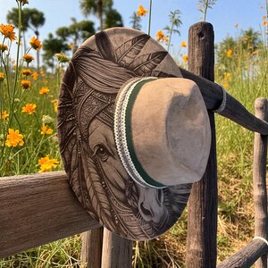 May include: A tan felt hat featuring a detailed dark brown laser-engraved illustration of a horse wearing a feathered headdress on the brim. The hat includes a dark green ribbon band accented with a thin white decorative trim around the crown.