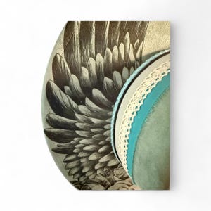 May include: A wide-brimmed hat featuring a detailed black and white illustration of large bird wings on the side. The hat is accented with a thin light blue cord, a white lace trim, and a wider band of solid teal fabric around the crown.