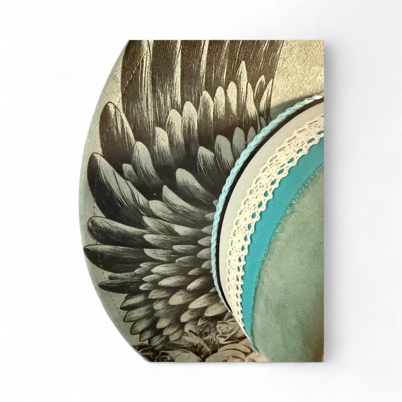 May include: A wide-brimmed hat featuring a detailed black and white illustration of large bird wings on the side. The hat is accented with a thin light blue cord, a white lace trim, and a wider band of solid teal fabric around the crown.