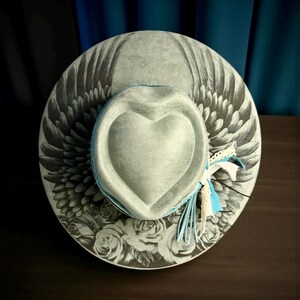 May include: A light gray wide-brimmed hat featuring a heart-shaped crown. The brim displays a black and white illustration of wings and roses. A light blue band encircles the base of the crown, accented with frayed white and blue fabric strips.