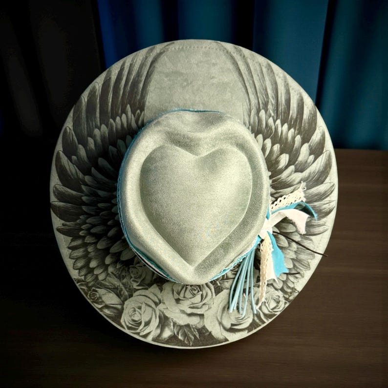 May include: A light gray wide-brimmed hat featuring a heart-shaped crown. The brim displays a black and white illustration of wings and roses. A light blue band encircles the base of the crown, accented with frayed white and blue fabric strips.