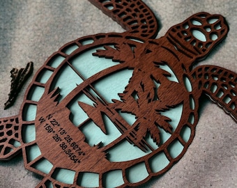 May include: A wooden turtle cutout with a light blue background. The turtle has a detailed design and the background has a palm tree silhouette. The turtle has coordinates inscribed on its shell: N 22° 13' 25.6874", W 159° 26' 38.3454".