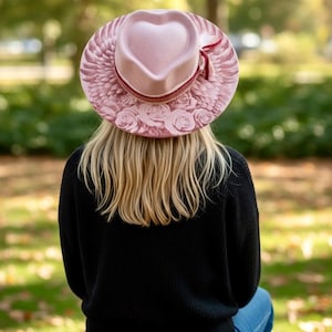 May include: A light pink hat with a heart-shaped crown, featuring a wide brim decorated with printed angel wings and rose patterns. The hat is accented with a thin, dark red ribbon band tied in a small bow on the side of the crown.