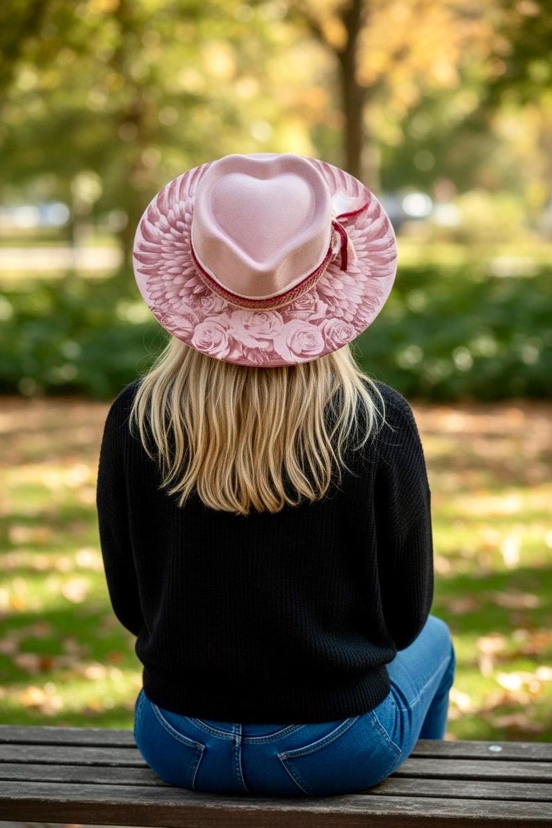 May include: A light pink hat with a heart-shaped crown, featuring a wide brim decorated with printed angel wings and rose patterns. The hat is accented with a thin, dark red ribbon band tied in a small bow on the side of the crown.