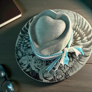 May include: A grey hat with a heart-shaped crown and a wide brim featuring a black and white print of roses and wings. The hat is accented with a layered trim of white lace and light blue ribbon tied in a bow on the side of the crown.