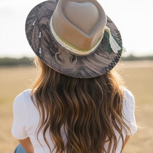 May include: A tan fedora hat with a heart-shaped crown, featuring a detailed black and white illustration of a horse on the brim. The hat is accented with a green ribbon, white lace trim, and dark stitching along the edge of the wide brim.