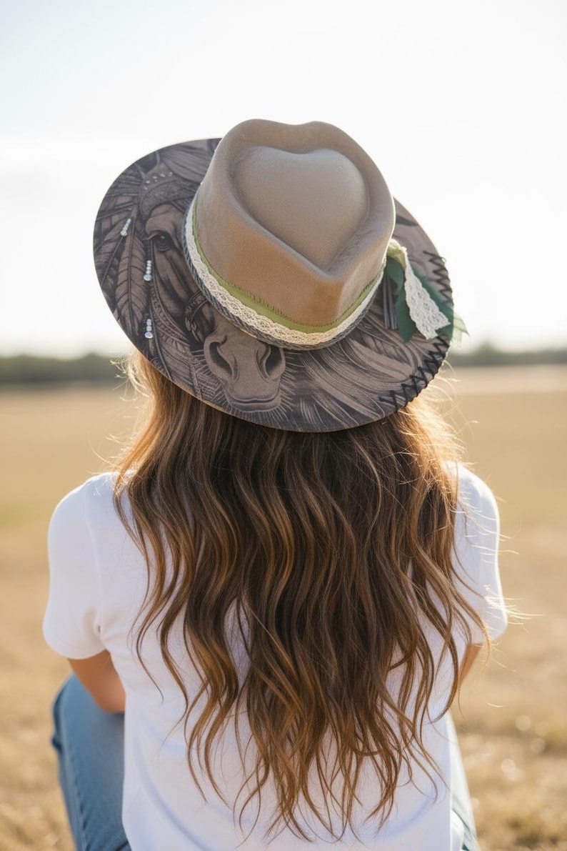 May include: A tan fedora hat with a heart-shaped crown, featuring a detailed black and white illustration of a horse on the brim. The hat is accented with a green ribbon, white lace trim, and dark stitching along the edge of the wide brim.