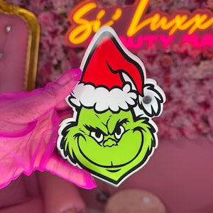 Może przedstawiać: Zielona ozdoba świąteczna z motywem Grincha z czerwoną czapką Świętego Mikołaja i białym wykończeniem. Twarz Grincha jest szczegółowa z psotnym uśmiechem i czarnymi konturami. Ozdoba jest trzymana na różowym kwiatowym tle.