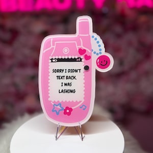 Puede incluir: Un letrero con forma de teléfono plegable rosa que presenta una pantalla blanca con el texto negro SORRY I DIDN'T TEXT BACK, I WAS LASHING. El letrero incluye corazones rosas, una estrella azul, una flor rosa, una nota musical y una cara sonriente.