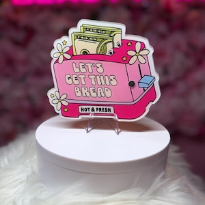 May include: A pink acrylic toaster-shaped sign featuring white flowers and yellow stars. The text reads LET'S GET THIS BREAD and HOT & FRESH. Two one-hundred-dollar bills are visible inside the toaster slots. The sign has a blue lever.