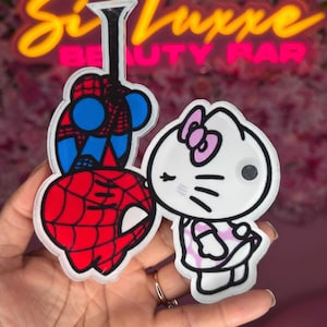 Op de afbeelding: Een rood en blauw Spiderman-personage en een wit Hello Kitty-personage met roze strikken en een roze jurk. De personages bevinden zich op een transparante achtergrond.
