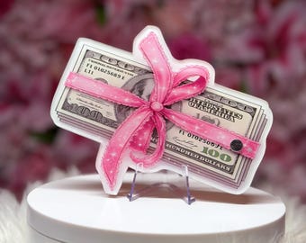 May include: A stack of United States one hundred dollar bills tied with a bright pink ribbon featuring a glittery star pattern. The ribbon is secured with a small black circular stud, and the entire money bundle is presented as a flat acrylic cutout.