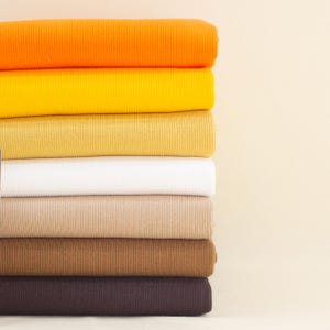 May include: A stack of folded fabric in various colors, including orange, yellow, white, beige, brown, and dark brown. The fabric has a ribbed texture, and the stack is positioned against a light beige background.