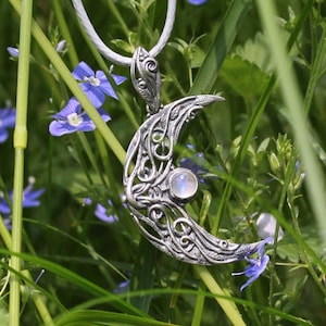 May include: A silver crescent moon pendant with a moonstone in the center, hanging from a white cord. The pendant is surrounded by green grass and blue flowers.