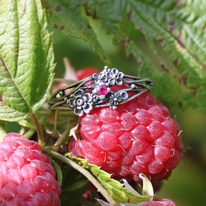 May include: A silver ring with a pink gemstone and floral accents, resting on a red raspberry. The ring is surrounded by green leaves.