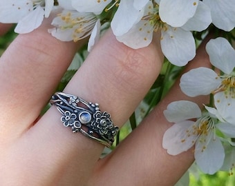 May include: A silver ring with a delicate floral design and a moonstone accent. The ring is worn on a hand holding a branch of white blossoms.
