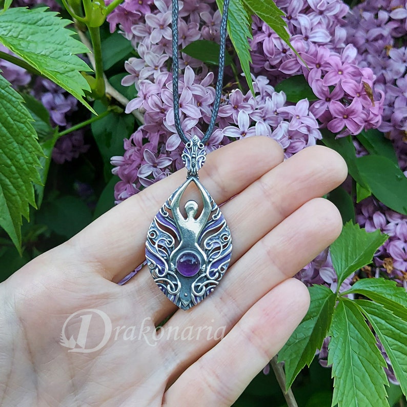 May include: Silver pendant necklace with a purple amethyst gemstone in the center. The pendant features a detailed design of a woman with outstretched arms, surrounded by intricate floral patterns.