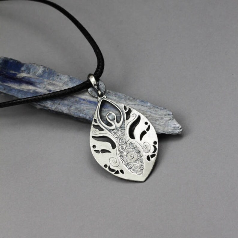 May include: A silver pendant necklace with a detailed design of a woman standing in a leaf shape. The pendant is on a black cord.