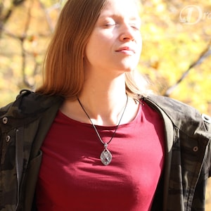 May include: A woman wearing a red shirt and a camouflage jacket with a silver pendant necklace. The pendant is a detailed design of a tree with leaves.