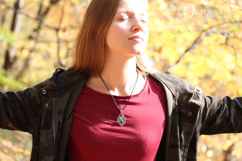 May include: A woman wearing a red shirt and a camouflage jacket with a silver pendant necklace. The pendant is a detailed design of a tree with leaves.