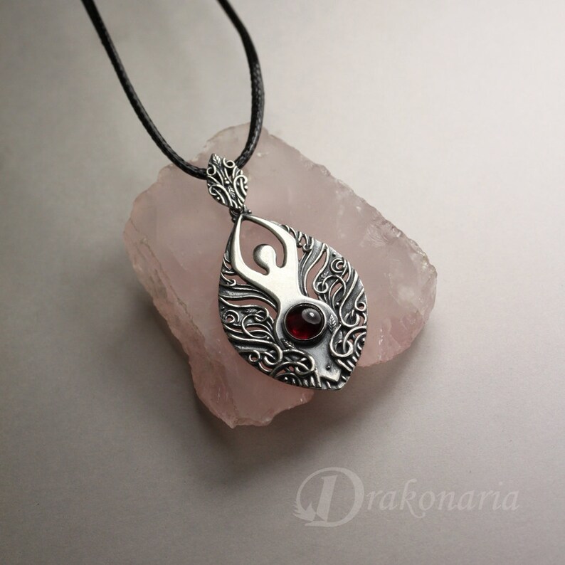 May include: A silver pendant necklace with a detailed design of a woman's figure with a red gemstone in the center. The pendant is hanging on a black cord.