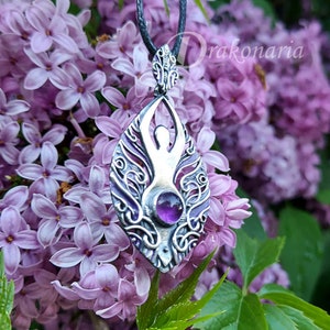 May include: A silver pendant featuring a stylized female figure with an amethyst gemstone in the center. The pendant is surrounded by intricate floral designs and hangs from a black cord.